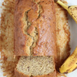 Loaf of banana bread, partially sliced on white marble, ripe banana on right.