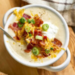 Potato soup topped with bacon, green onion, shredded cheese in white soup crock.