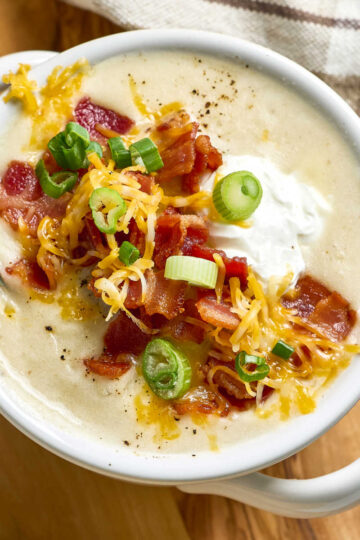 Potato soup topped with bacon, green onion, shredded cheese in white soup crock.