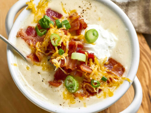 Potato soup topped with bacon, green onion, shredded cheese in white soup crock.