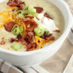 small white bowl of potato soup topped with crumble bacon, sour cream, shredded cheese, and sliced green onions