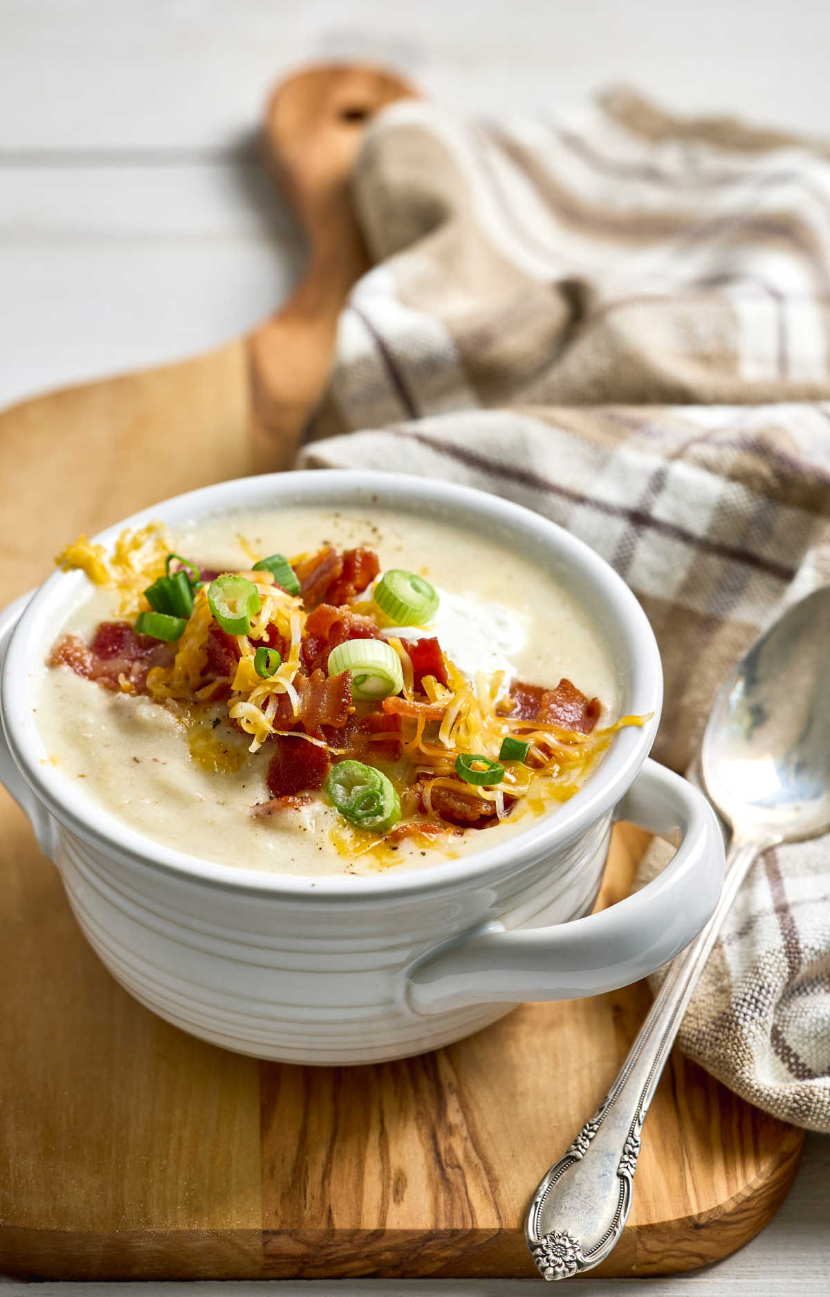 Potato soup in white soup crock topped with bacon, cheese, green onion.