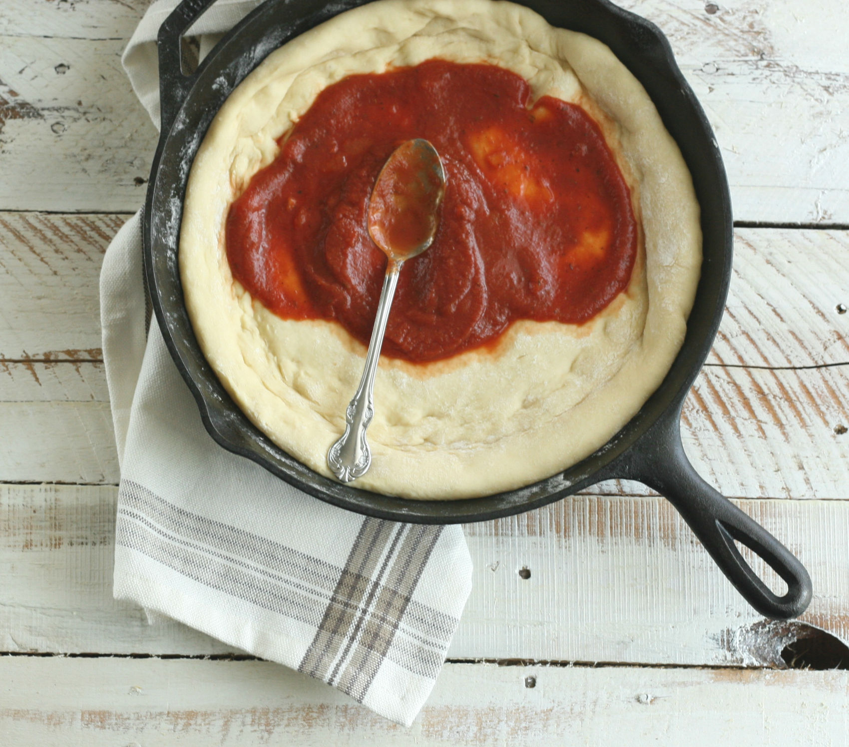 Pizza dough in cast iron skillet with spoon spreading marinara sauce.