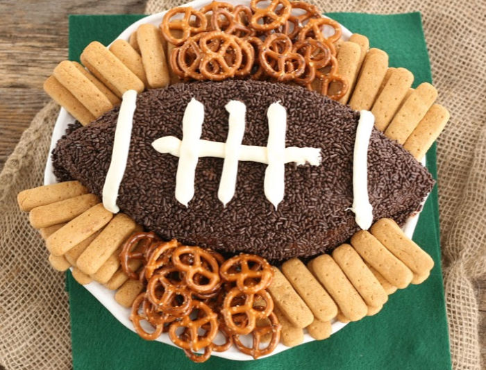 brownie batter dip shaped in a football, covered in chocolate sprinkles, graham cracker sticks and pretzels on plate