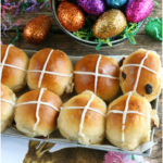 Hot cross buns with icing sitting in a galvanized tray and glittered Easter eggs to the background
