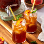 Glasses of tea with fresh peach slices, ice cubes and mint on wood cutting board.