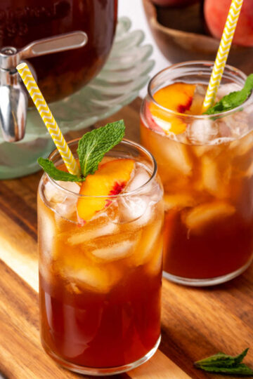 Glasses of tea with fresh peach slices, ice cubes and mint on wood cutting board.