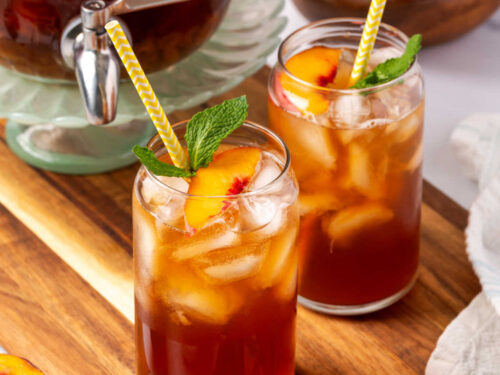 Glasses of tea with fresh peach slices, ice cubes and mint on wood cutting board.