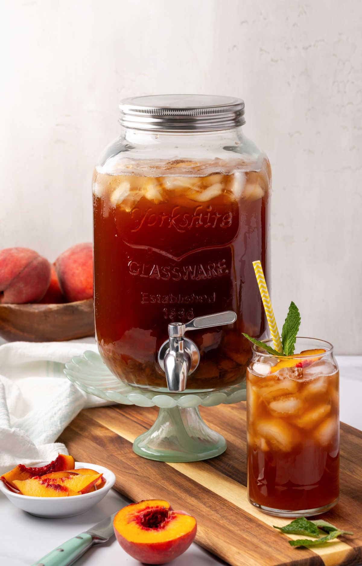 Glass drink dispenser with iced tea, glass of tea, ice cubes, fresh mint, peach slices in bowl.