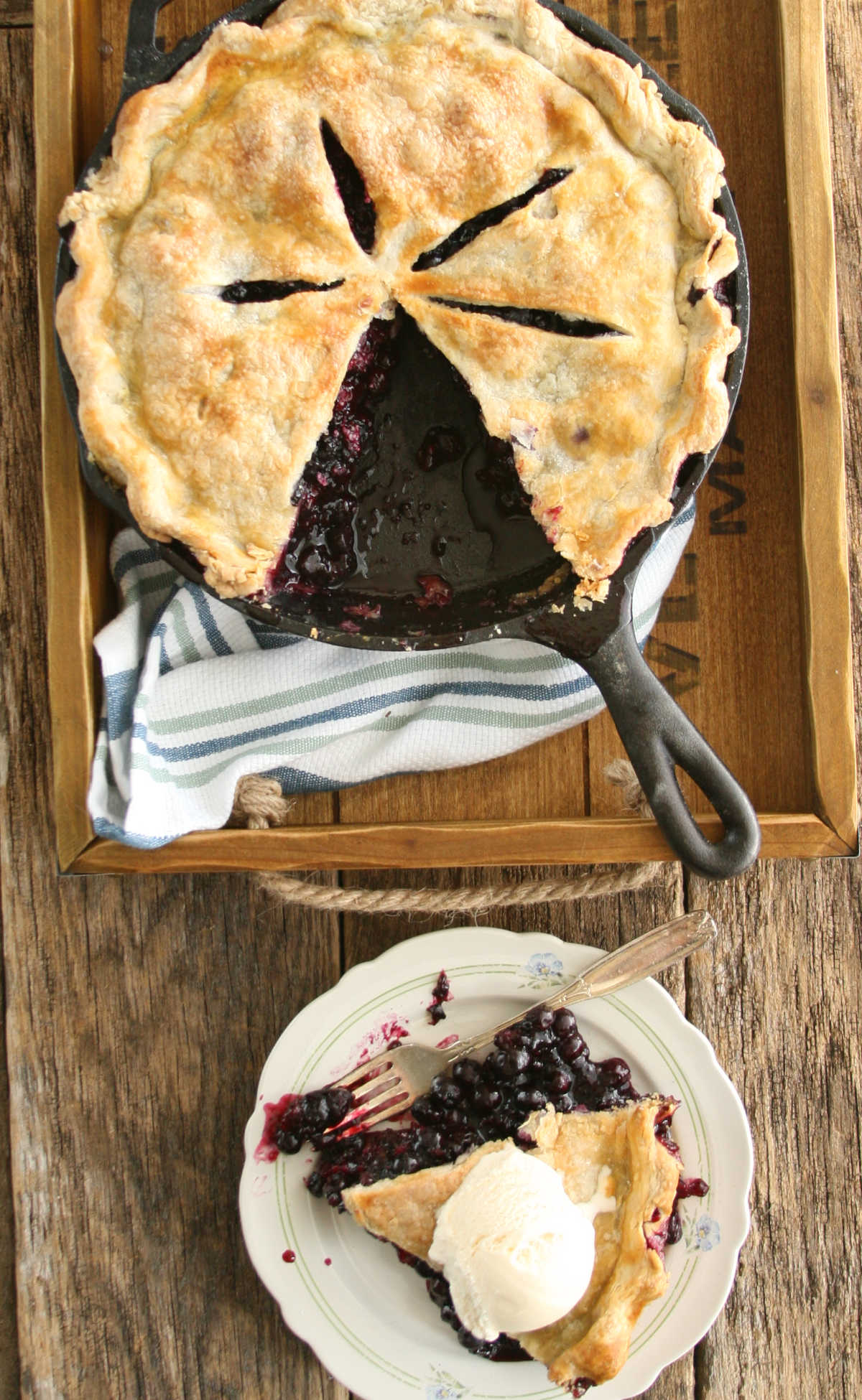 Blueberry pie in cast iron skillet on reclaimed wood boards, slice of pie on small white plate.
