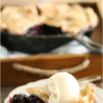 slice of blueberry pie with scoop of vanilla ice cream on white scalloped plate, blueberry pie in cast iron skillet in background