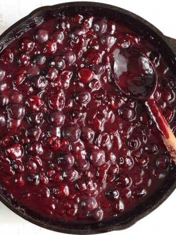 Cherry pie filling in large cast iron skillet with wooden spoon in pan, on white marble.