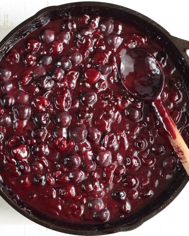 Cherry pie filling in large cast iron skillet with wooden spoon in pan, on white marble.