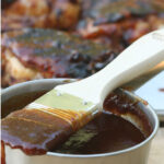 barbecue sauce in metal measuring cup, wooden handle pastry brush on top, dripping sauce, barbecue chicken in background