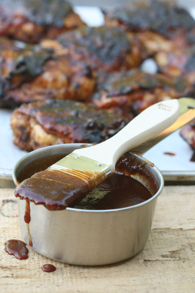 barbecue sauce in metal measuring cup and wooden handle brush on top dripping with barbecue sauce chicken in background