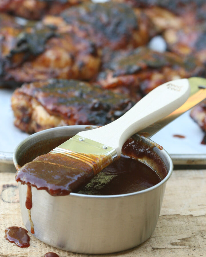 barbecue sauce in metal measuring cup and wooden handle brush on top dripping with barbecue sauce chicken in background