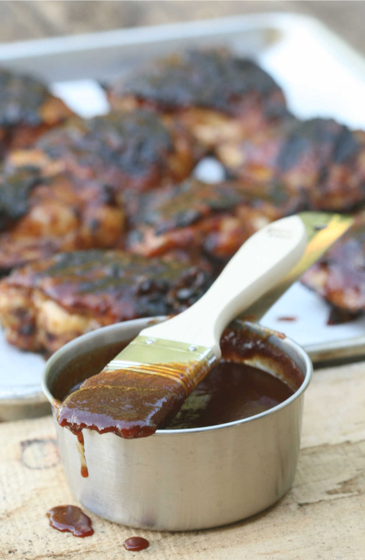 Pastry brush dripping with BBQ sauce on metal measuring cup, barbecue chicken in background.