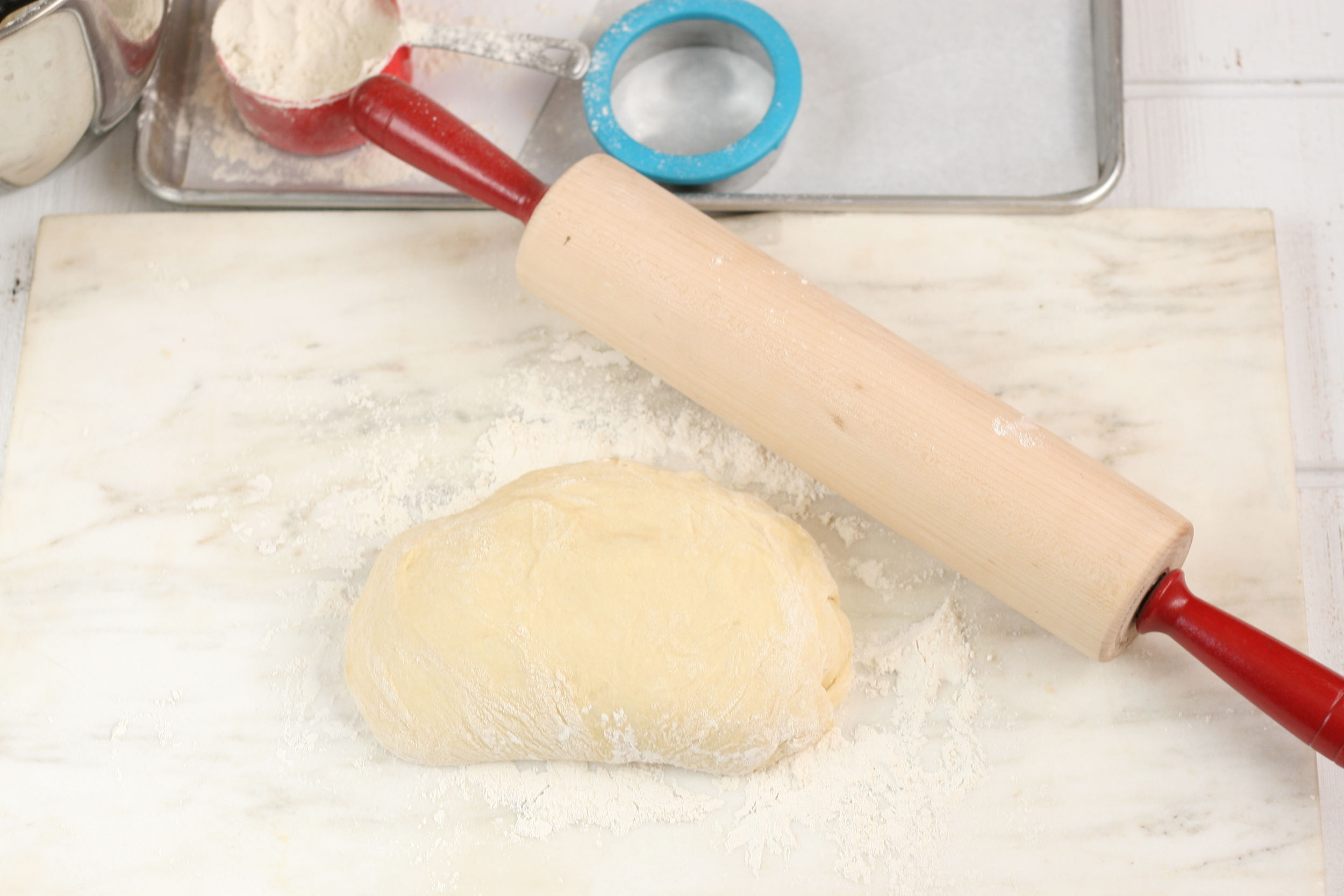 donut dough being rolled out on a piece of white marble and wooden rolling pin with red handles.