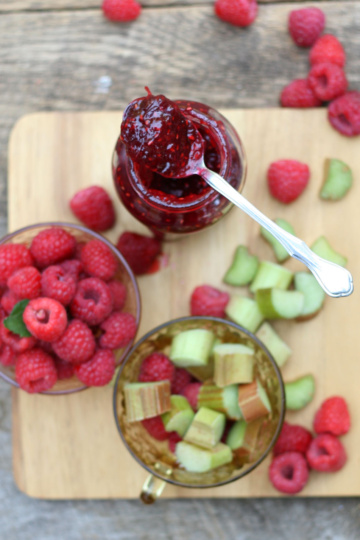 raspberry rhubarb jam on spoon on top of glass jar with loose berries and rhubarb on wooden cutting board