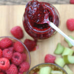 glass jar of raspberry rhubarb jam on wooden cutting board, slices of rhubarb, fresh raspberries surrounding.