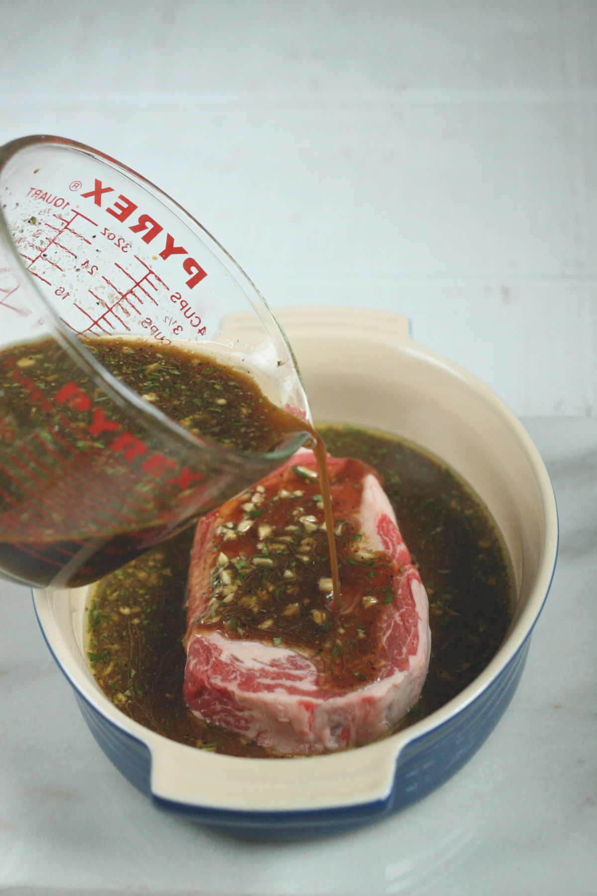 steak marinade for grilled steak in glass measuring cup being poured over NY strip steak in ceramic baking dish.