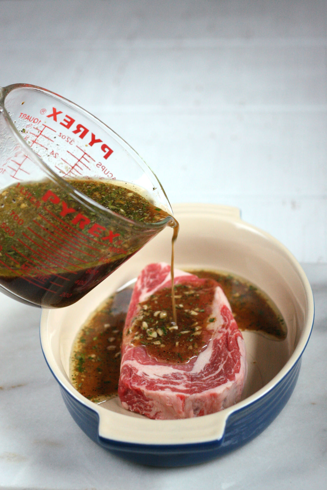 steak marinade being poured over a ribeye steak in an oval ceramic baking dish.