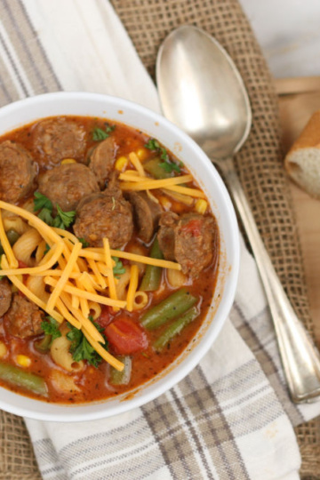 bowl of Italian sausage soup, topped with shredded yellow cheddar cheese and slices of French baguette to side of bowl