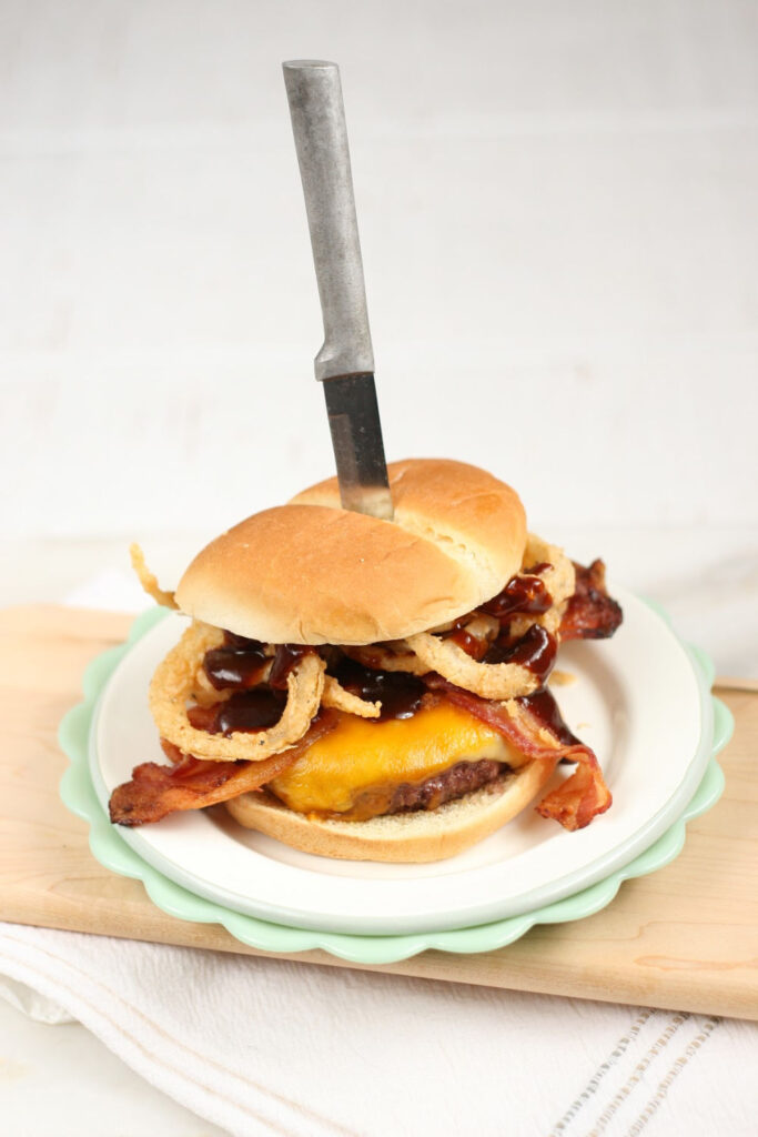 Bacon cheeseburger topped with buttermilk onion rings, cheddar cheese, and barbecue sauce on bun. Knife down center of burger