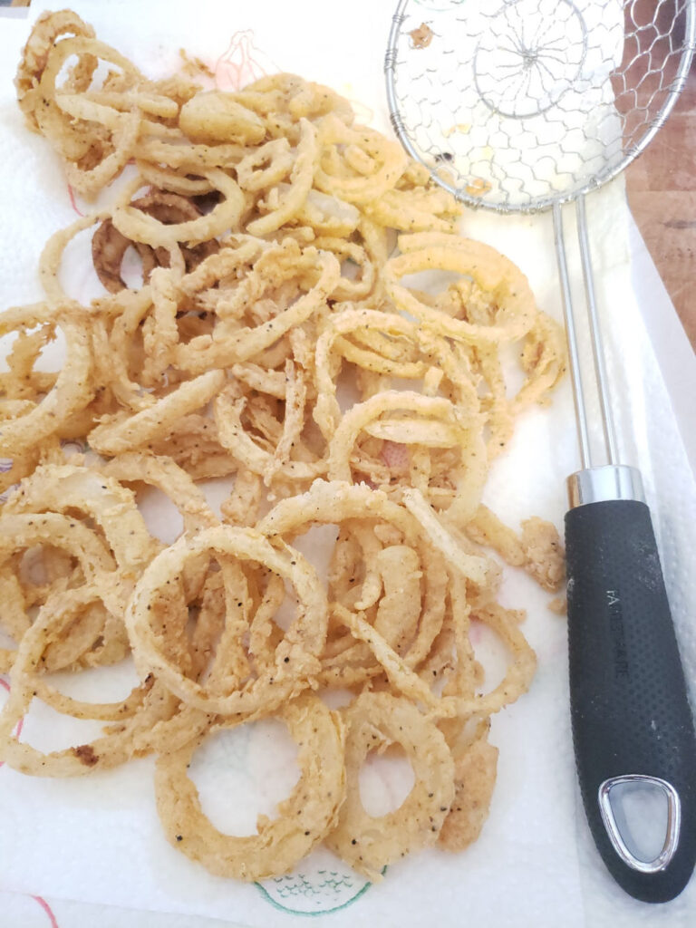 Buttermilk fried onion rings draining on paper towels on half sheet pan with strainer on right