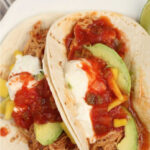 two soft tacos on small white plate with shredded chicken, salsa and avocado slices