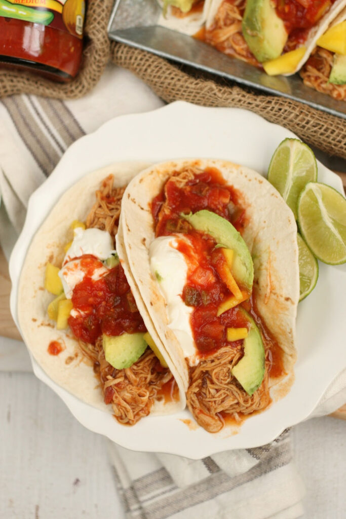 Mexican shredded chicken on soft taco shells with salsa, slices of avocado, and sour cream on small white plate