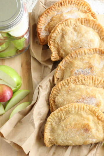 Fried apple hand pies on wooden cutting board, red and green apple quarters around.