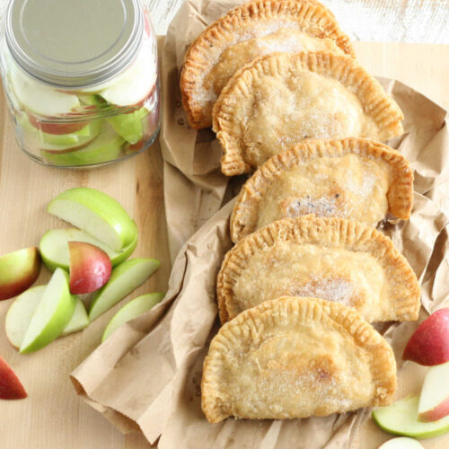 Fried apple hand pies on wooden cutting board, red and green apple quarters around.