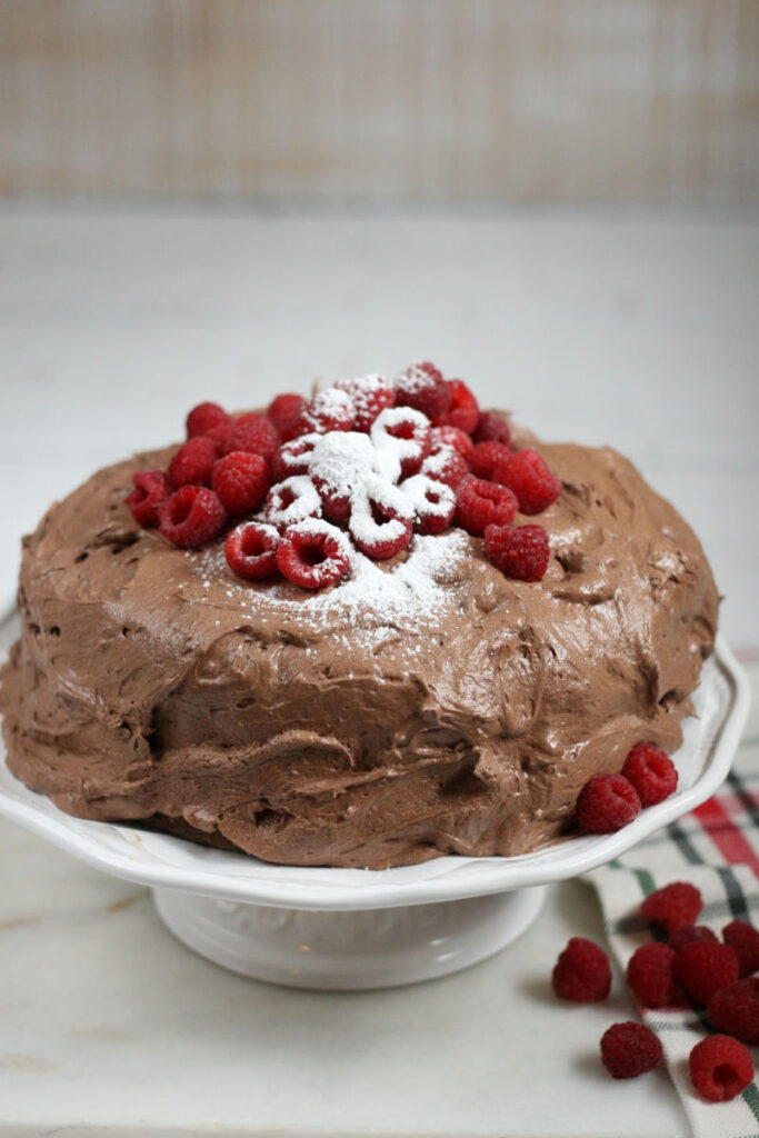 chocolate cake topped with fresh raspberries on a white footed cake dish