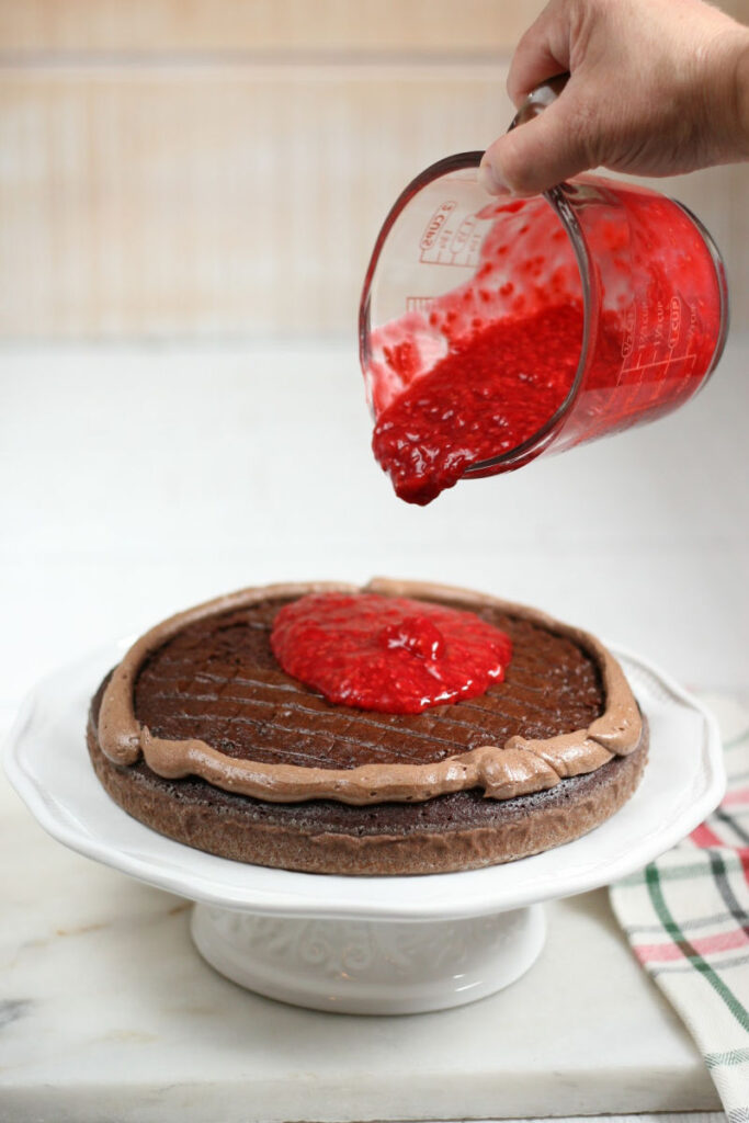 chocolate cake on white footed cake dish, pouring raspberry filling on top