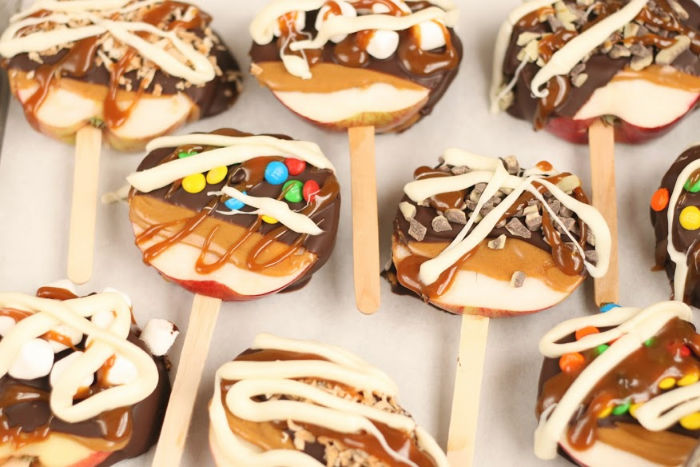 slices of apples on wooden sticks dipped in peanut butter and chocolate. topped with mini m&m's, toasted coconut, and white chocolate