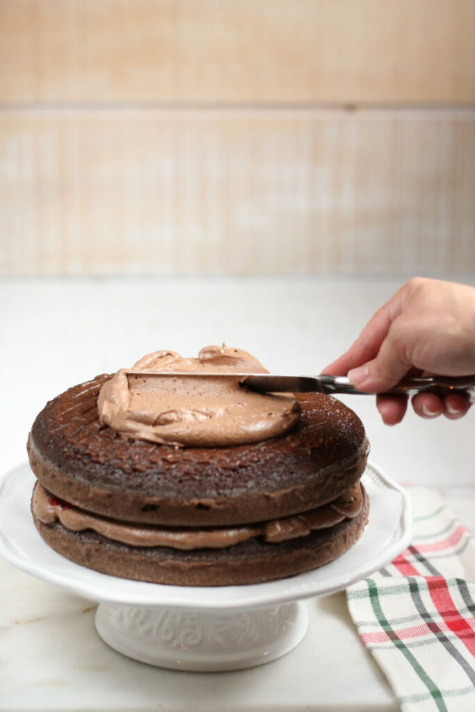 Chocolate layered cake on white footed cake dish spreading chocolate frosting with a butter knife on top of cake