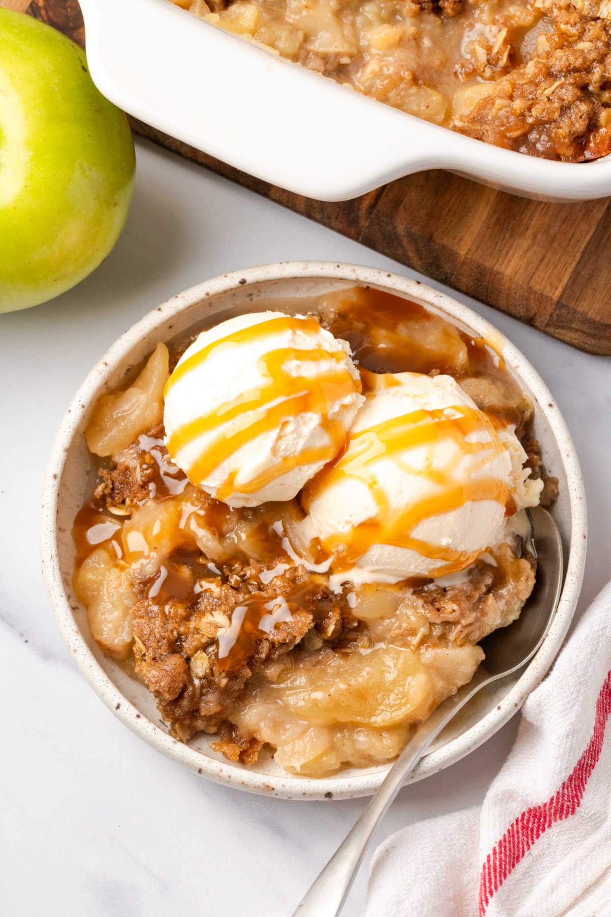 Apple crisp in white speckled bowl, topped with vanilla ice cream and caramel.