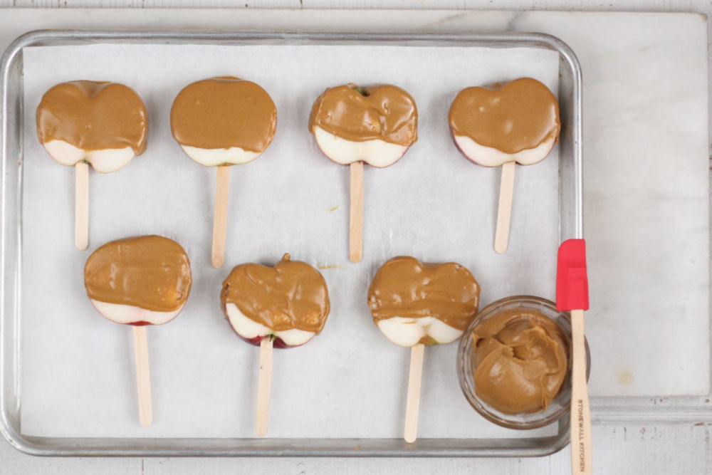 slices of apples with wooden sticks, dipped in peanut butter, setting on a half sheet pan