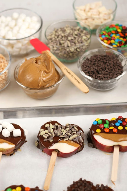 apple slices on wooden sticks, dipped in chocolate and toppings on half sheet pan. Various toppings in clear glass bowls