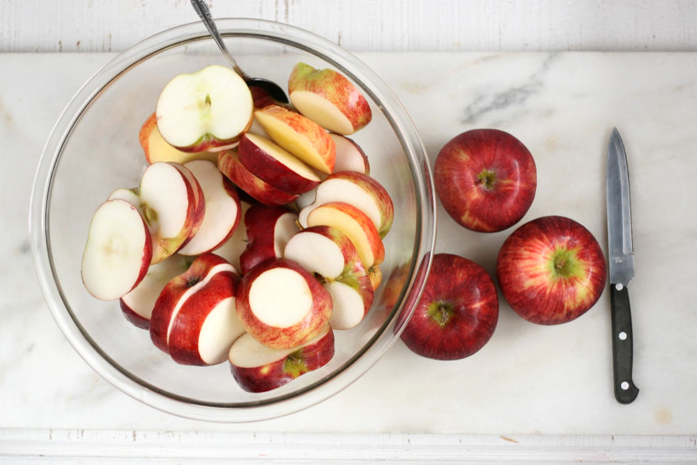 apple slices in a clear glass bowl on white marble with knife on right side