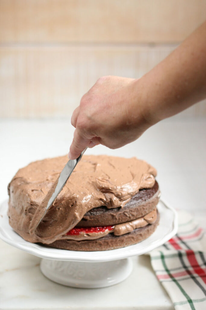 spreading chocolate frosting on chocolate layered cake on white footed cake dish