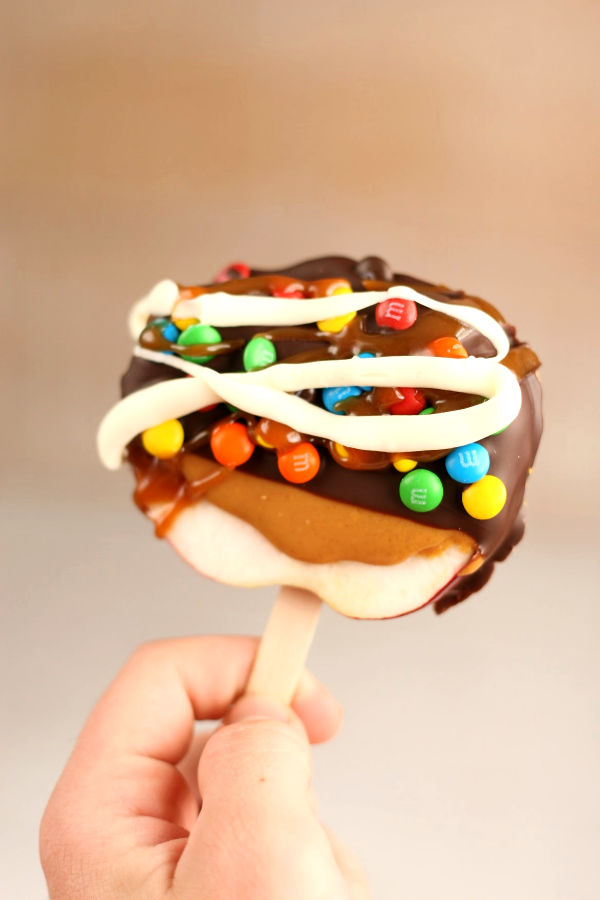 apple slice being held by hand, dipped in chocolate, mini m&m's, drizzled with white chocolate