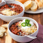 Chili in small white bowl topped with cheese, sour cream and jalapeno slices.