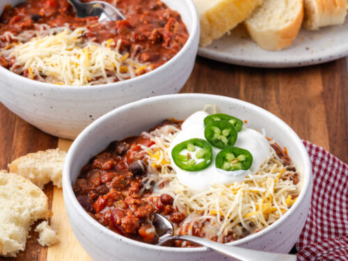 Chili in small white bowl topped with cheese, sour cream and jalapeno slices.