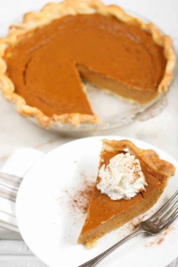 Pumpkin pie with slice cut out in glass pie dish, slice of pie on small white plate.
