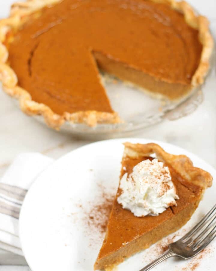 Pumpkin pie with slice cut out in glass pie dish, slice of pie on small white plate.