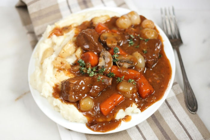 Beef stew with carrots, mushrooms, and pearl onions on top of mashed potatoes on white plate, fork to the right