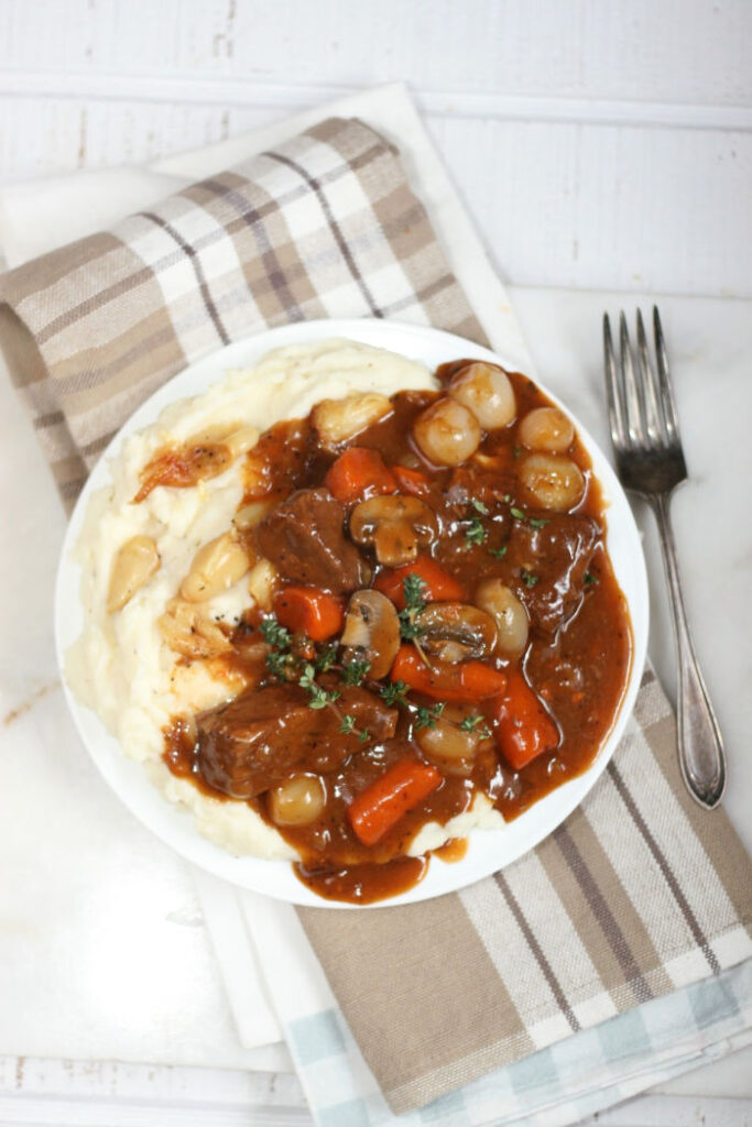 boeuf bourguignon on a white plate with mashed potatoes. Sitting on taupe plaid napkin with fork to right