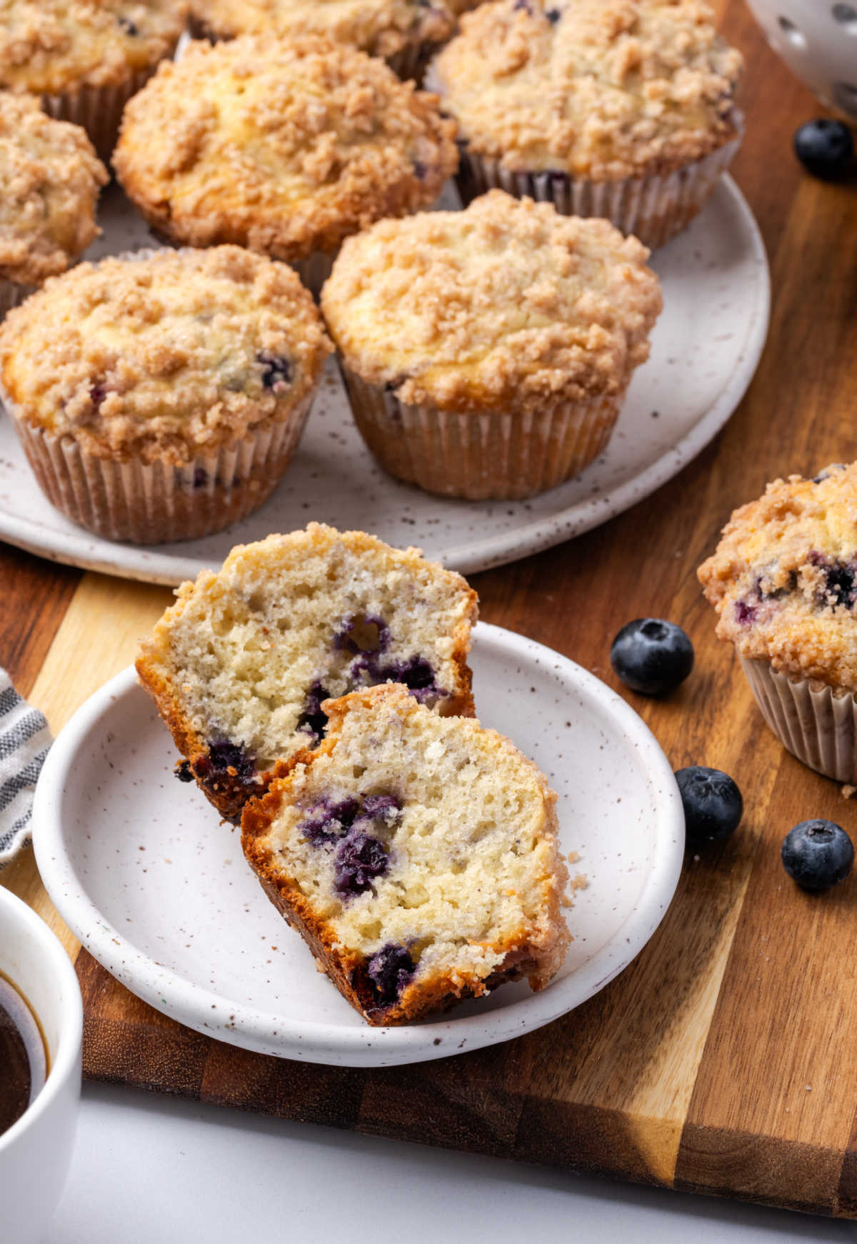 Blueberry muffin cut in half on small white plate, more muffins in background.